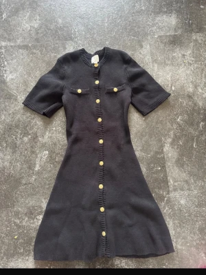 Svart stickad klänning med guldknappar - Snygg svart stickad klänning med korta ärmar och dekorativa guldknappar framtill. Klänningen har en figurnära passform och två fejkade bröstfickor. Perfekt för dig som gillar klassisk men trendig stil.