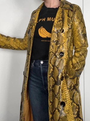 STAND STUDIO  - Gul trenchcoat med ormmönster - Cool trenchcoat från Stand studio i gul och svart med ormmönster och glansig finish. Dubbelknäppt med svarta knappar och klassisk krage. Storlek S/M. 