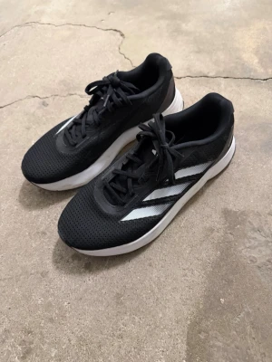 Svarta Adidas duramo  - Säljer ett par svarta Adidas sneakers med klassiska vita ränder på sidan. Skorna har en mesh-överdel som ger bra ventilation och en vit, tjock sula för komfort. Perfekta för dig som gillar sportig stil och vill ha något som funkar till jeans eller träningskläder.