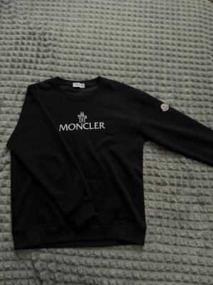Svart Moncler sweatshirt med logga - Cool svart sweatshirt från Moncler med vit logga på bröstet och Moncler-märke på ärmen. Tröjan har rund hals och ribbade muddar. Perfekt för dig som gillar stilrena och exklusiva plagg.