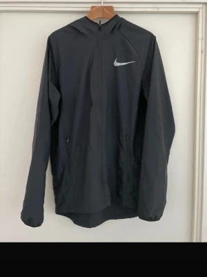 Svart Nike vindjacka med huva - Säljer en svart vindjacka från Nike med huva och hel dragkedja. Jackan har två fickor med dragkedja och ett stort vitt Nike-logga på bröstet. Materialet är lätt och perfekt för löpning eller sport. Stilen är enkel och sportig.