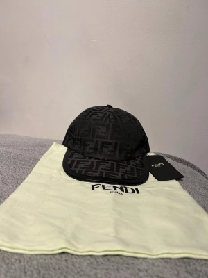 Svart Fendi keps med FF-mönster - Säljer en svart keps från Fendi med det klassiska FF-logomönstret över hela kepsen. Kepsen har böjd skärm, justerbart band bak och är tillverkad i ett tygmaterial. Perfekt för dig som vill ha en stilren och lyxig accessoar med tydlig logga.