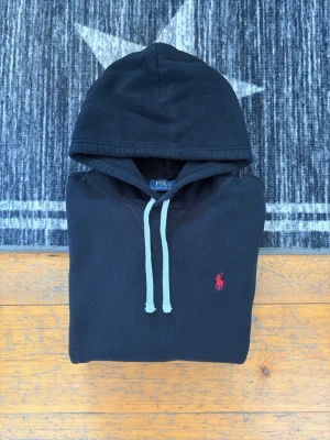 Svart Polo Ralph Lauren Fleece Hoodie - Storlek M - Hej! 👋 Säljer en klassisk svart Polo Ralph Lauren hoodie i storlek M. Den har den ikoniska röda pony-loggan broderad på bröstet – den mest tidlösa färgkombinationen! 🐎 Tröjan är i mycket fint skick, djupsvart i färgen och har den mjuka fleece-insidan kvar. Inga fläckar, inga hål och minimalt med noppor. Denna modell har även QR-kod för enkel verifiering av äkthet, vilket gör det tryggt och smidigt för dig som köpare ✅ Ett premium-basplagg som passar till allt. Fraktar snabbt! 🚛📦💬💯 