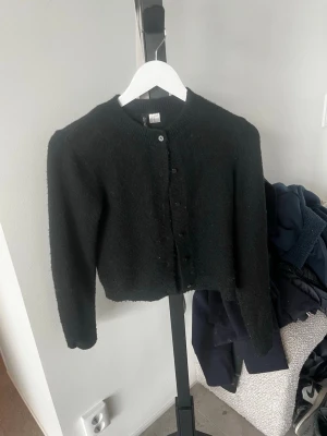 Svart kofta från Divided XS - Snygg svart kofta från Divided i storlek XS. Koftan har rund halsringning, knäppning framtill med svarta knappar och ribbade muddar vid ärmslut och nederkant. Perfekt att slänga över en topp för en enkel och stilren look.