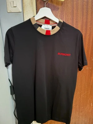 Burberry t-shirt oanvänd, Storlek S - Cool svart t-shirt från Burberry med röd logga på bröstet och unik krage i beige, svart och röd. Perfekt för dig som vill ha något stilrent men ändå sticker ut. Klassisk passform och mjukt bomullsmaterial som känns skönt hela dagen.