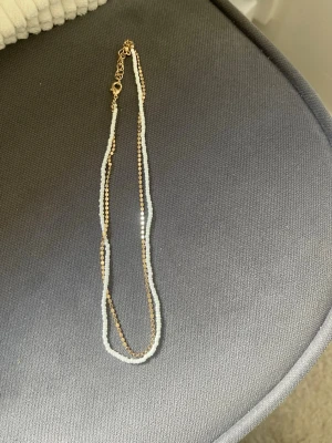 Tvådelat halsband med pärlor och gulddetaljer - Säljer ett snyggt halsband med två separata kedjor, en med vita pärlor och en med små guldfärgade pärlor. Halsbandet har guldfärgad kedja och justerbar låsning. Perfekt för att ge din outfit en cool touch.