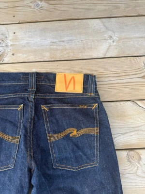 Mörkblå jeans från Nudie Jeans - Snygga mörkblå jeans från Nudie Jeans med klassisk rak passform. Jeansen har tydliga orangea sömmar och Nudie-loggan på bakfickan samt en läderpatch i midjan. Tillverkade i denim av bomull.