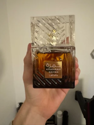 Lattafa Khamrah Qahwa parfym - Lattafa Khamrah Qahwa är en parfym i en lyxig, fyrkantig glasflaska med mönstrad design. Flaskan har en gradient från mörkbrun till klar och ett elegant guldmärke framtill. Locket är transparent med snidade detaljer som ger en exklusiv känsla.