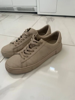 Beige sneakers i läder-look - Snygga beige sneakers med klassisk låg profil och rund tå. Skorna har en stilren design med matchande beige snörning och sula. Materialet ser ut att vara läder eller läderimitation, vilket ger en clean och modern vibe. Perfekt för dig som gillar minimalistisk stil.
