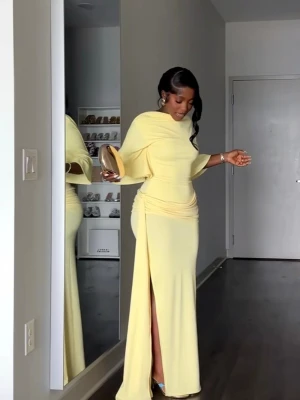 Ljusgul långklänning med slits - Ljusgul långklänning med elegant slits framtill och öppen rygg. Klänningen har breda, flödande ärmar och draperad midja som ger en snygg siluett. Perfekt för dig som vill ha en unik och stilren look till sommaren. 