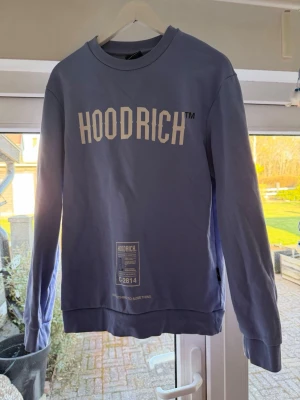Blå HOODRICH sweatshirt med tryck - Cool blå sweatshirt från HOODRICH med stort vitt tryck på bröstet och detaljerad grafisk text på ryggen. Tröjan har rund hals, långa ärmar och ribbade muddar. Perfekt för dig som gillar streetwear och vill sticka ut.