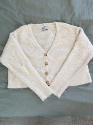 Vit fluffig kofta med knappar - Mysig vit kofta från Bershka med fluffig känsla och v-ringning. Koftan har långa ärmar och fyra beige knappar framtill. Perfekt för lager på lager och ger en trendig look till din outfit.