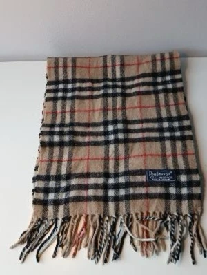Beige rutig halsduk från Burberry - Klassisk beige halsduk från Burberry med det ikoniska rutmönstret i svart, vitt och rött. Halsduken är tillverkad i 100% lammull och har fransar i ändarna. Perfekt accessoar för att lyfta din outfit med en tidlös brittisk stil.