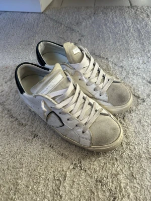 Philippe Model vita sneakers storlek 39 - Snygga vita sneakers från Philippe Model med svart detalj på hälen och sidan. Skorna har klassisk låg profil, vita skosnören och en blandning av läder och mocka på ovandelen. Perfekta för dig som gillar stilrena och trendiga sneakers. Storlek 39. 