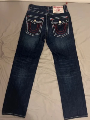 True Religion mörkblå jeans - Snygga mörkblå jeans från True Religion med kontrasterande vit och röd söm. Klassisk femficksdesign och stora bakfickor med lock och knappar. Jeansen har raka ben och är tillverkade i denim. Perfekt för dig som gillar detaljer och ikoniska märken.