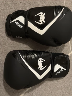 Svarta boxningshandskar från Venum - Säljer ett par svarta boxningshandskar från Venum med vita detaljer och logga. Handskarna har kardborreknäppning vid handleden för bra passform och är tillverkade i slitstarkt syntetmaterial. Perfekta för boxning eller träning på gymmet.  Diskuterar pris privat. 14 prisförslag men det fortfarande säljs. Köp nu finns inte bättre pris än de här.