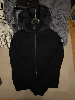 Moncler cardigan  - Svart Moncler cardigan i nyskick med stickad utsida och vadderad insida. Jackan har dragkedja framtill, stor huva och ribbstickade muddar. frågor eller funderingar tveka inte på att höra av dig