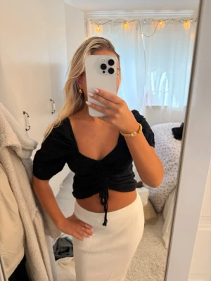 Svart croppad topp med puffärm - Trendig svart croppad topp med puffärmar och djup v-ringning. Toppen har en snygg dragsko framtill som ger en cool rynkad effekt. Perfekt till höga kjolar eller jeans för en modern look.