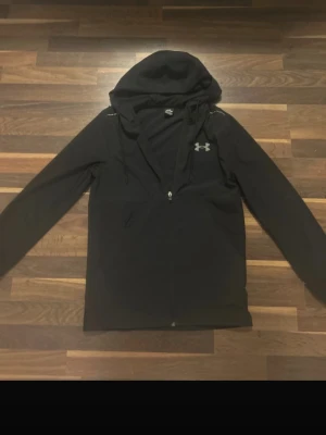Svart hoodie från Under Armour - Snygg svart hoodie från Under Armour med dragkedja framtill och justerbar huva. Tillverkad i ett lätt och ventilerande material med diskret logga på bröstet. Perfekt för dig som gillar sportig och clean stil.