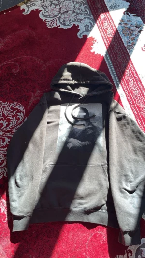 Svart hoodie med Mona Lisa-tryck - Säljer en svart hoodie med stor huva och en unik Mona Lisa-print på bröstet. Hoodien har klassisk känguruficka framtill och ribbade muddar vid ärmar och nederkant. Perfekt för dig som gillar streetwear och konstinspirerade plagg.