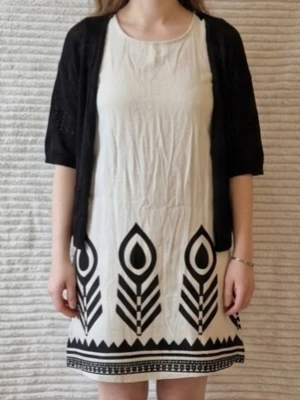 Boho-set: Mönstrad tunika & tunn stickad kofta stl M - ​Mönstrad ärmlös tunika/klänning: En ljus, krämvit tunika i ett skönt naturmaterial med ett iögonfallande svart grafiskt mönster (fjäder/blad-motiv) nedtill. Snygg till både jeans, leggings eller bara ben. ​Svart tunnstickad kofta (Primark): En nätt, svart kofta med snygga hålstickade detaljer som ger en lätt och luftig känsla. Perfekt att slänga över tunikan när det blir lite kyligare.