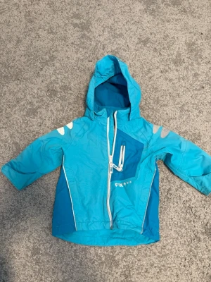 Blå skaljacka från FIX med huva - Snygg blå vindjacka från FIX med dragkedja, huva och reflexdetaljer på axlarna. Jackan har en sned bröstficka med vit dragkedja och vita detaljer längs sömmarna. Tillverkad i lätt och tåligt material, perfekt för blåsiga dagar.