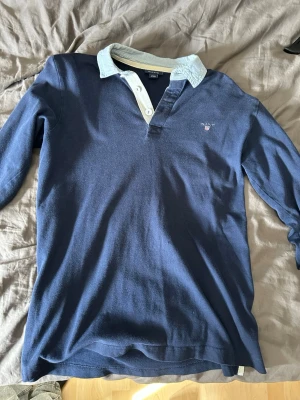 Blå långärmad henley från Gant - Snygg blå långärmad henley från Gant med ljusblå krage och vita knappar. Skjortan har broderad Gant-logga på bröstet och är tillverkad i mjuk bomull. Perfekt för en avslappnad men stilren look. Storlek 170. Ett jätte litet hål som man inte ser.