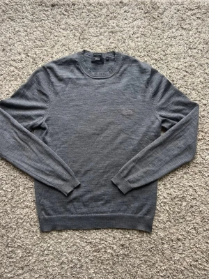 Grå stickad tröja från BOSS - Stilren grå stickad tröja från Hugo BOSS med rund halsringning och diskret broderad logga på bröstet. Perfekt för lager på lager och enkel att matcha. Snyggt med skjorta under. (Personlig favorit)