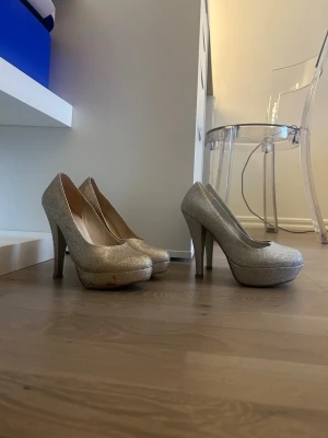 Glittriga silverfärgade och guldfärgade klackskor  - Säljer ett par snygga klackskor i silver och guld med glittereffekt och hög platåklack. Skorna har rundad tå och klassisk siluett, perfekta för dig som vill sticka ut. Materialet är syntet med glittrig yta. De båda tillsammans säljs för 120kr