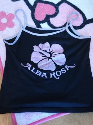 Svart linne Alba Rosa med tryck - Snyggt svart linne från Alba Rosa med tunna axelband och ljuslila kant (Brukade vara ljus rosa) Framtill finns ett stort rosa hibiskusblom-tryck och texten 'ALBA ROSA'. Perfekt till sommaren och lätt att matcha med jeans eller kjol. Finns liten sprickning av sömnaden men går stt fixa. Priset kan diskuteras om! 💵💸😋 