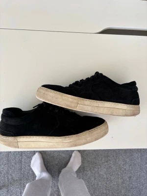 Svarta sneakers från Axel arigato  - Stilrena svarta sneakers från Axel arigato i mocka med vit, rejäl gummisula. Klassisk låg modell med snörning och diskret design som passar till det mesta. Perfekta för dig som gillar en clean och enkel look.