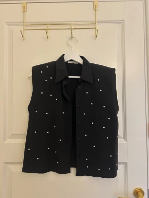 Svart väst med pärldetaljer från qlis  - Stilren svart väst från qlis med klassisk krage och knäppning framtill. Västen är dekorerad med små vita pärlor över hela framsidan för en trendig look. Perfekt att styla över en skjorta eller t-shirt för extra edge.