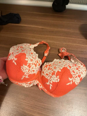 Orange push-up bh med spetsdetaljer - Snygg orange push-up bh med vadderade kupor och vacker vit spetsbrodyr över kuporna. Justerbara axelband och hakknäppning bak. Materialet är mjukt och stretchigt för extra komfort. Perfekt för dig som vill ha något färgglatt och elegant under kläderna.