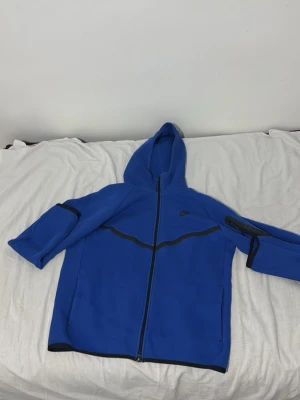 Blå Nike hoodie med dragkedja - Snygg blå hoodie från Nike med hel dragkedja framtill och svarta detaljer längs sömmarna. Utrustad med huva och fickor på sidorna. Perfekt för en sportig och avslappnad stil. Nike-logga på bröstet och cool design på ärmarna.