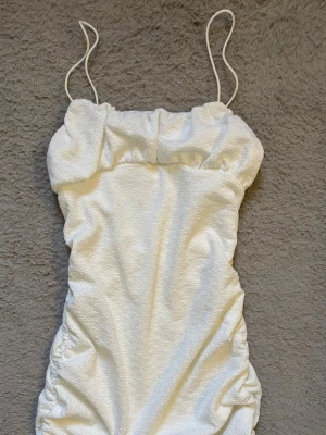 Vit kort klänning med tunna band - Trendig vit kortklänning med tunna axelband och rynkade sidor. Klänningen har en slät, lätt glansig yta och figurnära passform som ger en snygg siluett. Perfekt för sommarens alla tillfällen!