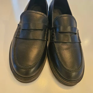 Svarta klassiska loafers i skinn - Säljer ett par stilrena svarta loafers i äkta skinn med klassisk penny loafer-design. Skorna har rundad tå, låg klack och diskreta sömmar längs kanterna. Perfekta för dig som gillar tidlös och enkel stil. Det står stl 37 i dem men det stämmer inte. De är 38, 5.