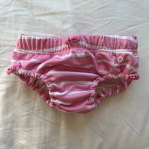 Rosa randiga badbyxor POP 62/68 - Säljer ett par söta rosa och vita randiga badbyxor från Polarn O. Pyret. De har justerbara snoddar i midjan och vid benen för bästa passform. Perfekta för strand eller pool! Märkta med POP-logga fram.