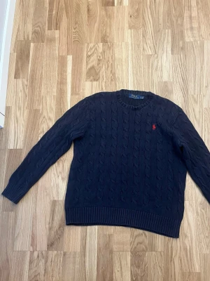 Mörkblå stickad tröja från Polo Ralph Lauren - Kabelstickad mörkblå tröja från Polo Ralph Lauren. Tröjan har rund halsringning, långa ärmar och ribbade muddar vid ärmslut och nederkant. Är insydd vid halsen men inget som syns