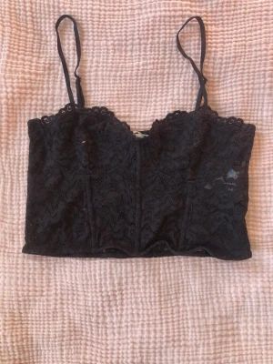 Svart spetsig croppad linnetopp - Säljer en svart croppad linnetopp från Gina Tricot med tunna axelband och vacker spets över hela toppen. Toppen har en feminin och elegant look med genombruten spets och lätt vågig kant nedtill. Perfekt att styla till jeans eller kjol.