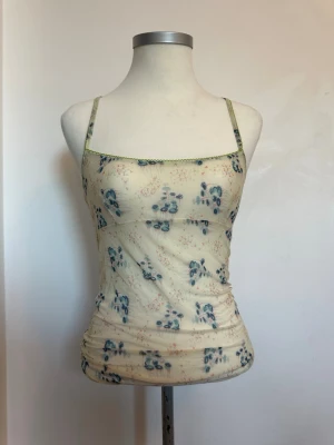 Blommig mesh linnetopp med smala band från Diesel - Söt linnetopp i mesh med blommigt mönster i blått och rosa på beige botten. Toppen har smala axelband och gröna sömmar längs kanterna. Figurnära passform och lätt transparent material, perfekt för varma dagar.