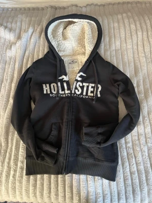 Svart Hollister hoodie med teddyfoder - Mysig svart hoodie från Hollister med dragkedja och stor vit logga på bröstet. Insidan är fodrad med fluffigt vitt teddyfoder, även i huvan. Två fickor framtill och ribbade muddar vid ärmar och nederkant. Perfekt för kalla dagar och riktigt skön att ha på sig.