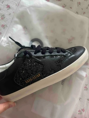 Svarta golden goose sneakers med glitter - TRYCK INTE KÖP NU!!! Kollar vad folk kan tänka sig betala för dessa  svarta sneakers från Golden Goose. Har glittriga detaljer på sidor och tå. Skorna har svarta skosnören, vit sula och GGDB/SSTAR-logga i guld på sidan. Materialet är en mix av läder och textil med glitterinslag för extra edge. Är i storlek 42 men passar som en 41, är 26,5cm i längd