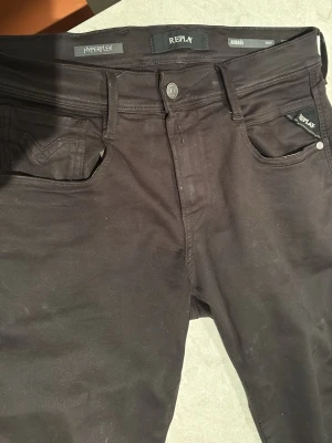 Svarta Replay Hyperflex jeans - Säljer ett par svarta Replay Hyperflex jeans med klassisk femficksdesign och snygga detaljer på bakfickorna. Jeansen har en slim fit passform och är tillverkade i ett stretchigt material som ger extra komfort. Perfekta för dig som gillar stilrena och bekväma jeans.