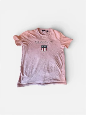 Ljusrosa GANT t-shirt med tryck - Säljer en ljusrosa t-shirt från GANT med klassiskt tryck framtill och ett emblem med stjärnor och ränder. T-shirten har rund hals och är tillverkad i mjuk bomull. Perfekt för dig som gillar stilrena och sportiga plagg.