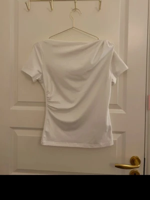 Vit omlottopp med drapering - Snygg vit topp med omlott-detalj och drapering på sidan. Kortärmad och figurnära passform som ger en modern och stilren look. Perfekt till jeans eller kjol för en trendig outfit.