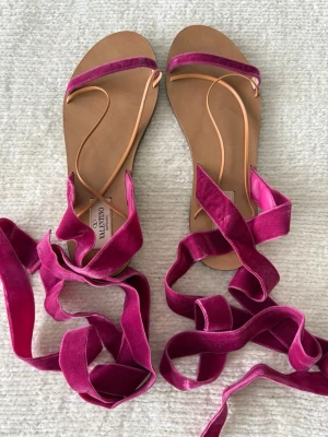 Rosa sammetsandaler från Valentino - Säljer ett par trendiga sandaler från Valentino med breda sammetsband i stark rosa och tunna beigea remmar. Sandalerna har platt sula och långa band som kan knytas runt ankeln för en cool look. Perfekta för att lyfta vilken outfit som helst.