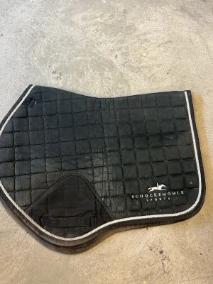 Svart schabrak från Schockemöhle - Säljer ett svart schabrak från Schockemöhle Sports med rutmönster och vit kant. Perfekt till ridning och har en klassisk design med broderad logga. Materialet är quiltat tyg och det finns förstärkning vid sadelgjorden. Är lortigt men blir rent av en tvätt