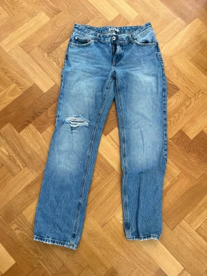 Blå raka jeans med hål från Only - Säljer ett par blå raka jeans från Only med slitning och hål på vänster lår. Jeansen har klassisk femficksmodell, normal midja och knappgylf. Materialet är denim i bomull och passformen är avslappnad och trendig.