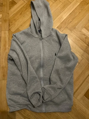 Grå zip hoodie från Polo Ralph Lauren - Snygg grå hoodie med dragkedja från Polo Ralph Lauren. Klassisk design med huva, fickor framtill och den ikoniska svarta loggan broderad på bröstet. Tillverkad i mjuk bomullsblandning som är skön att ha på sig. Perfekt för en avslappnad streetwear-look.