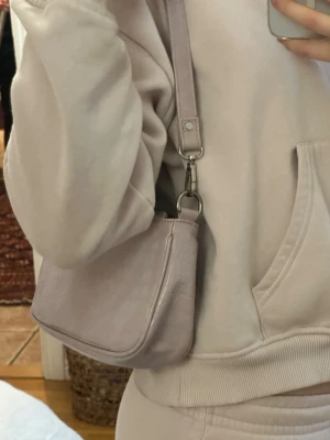 Ljuslila axelväska i croc-mönster - Säljer en ljuslila axelväska med croc-mönstrad yta och justerbar axelrem. Väskan har en stilren, rektangulär form och stängs med dragkedja. Perfekt för dig som vill ha en trendig och enkel accessoar till din outfit.